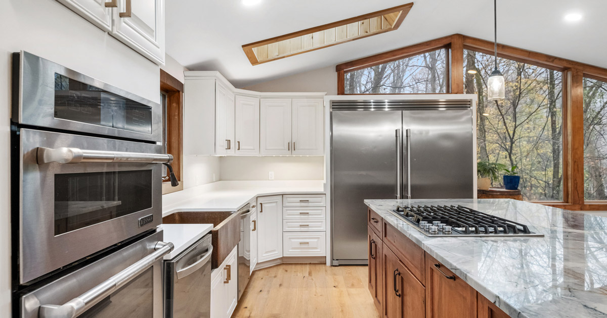 Modern kitchen with white cabinets, marble island with gas stove, stainless steel double oven and refrigerator, and large windows showing trees outside.