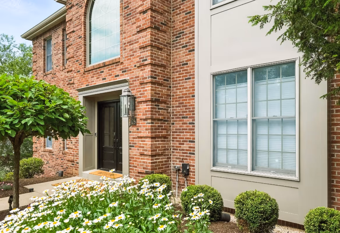 Front entrance of a brick house with black doors, an arched window above, and a garden with white daisies and trimmed bushes.