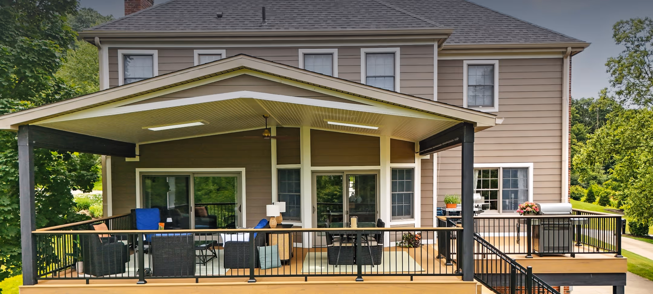 Covered outdoor patio with black railing, furnished seating area, dining table, and a grill attached to a two-story house with beige siding.