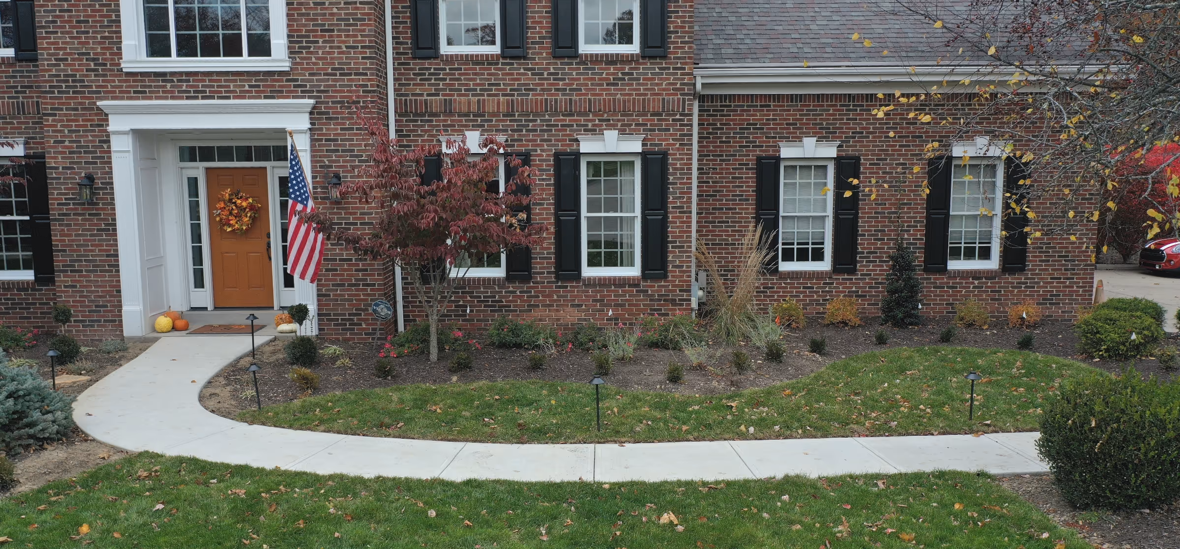 Front view of a brick house with an orange door decorated with a fall wreath, an American flag, and a curved concrete walkway through a landscaped garden bed and green lawn.