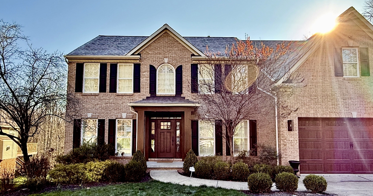 Two-story brick house with black shutters, a central wooden front door, surrounding bushes, and bright sunlight in the top right corner.