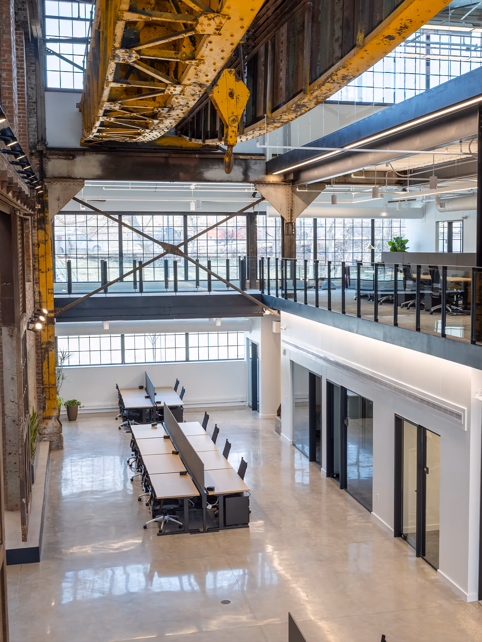 Modern industrial office space with long desks, ergonomic chairs, exposed yellow steel beams, and large windows.