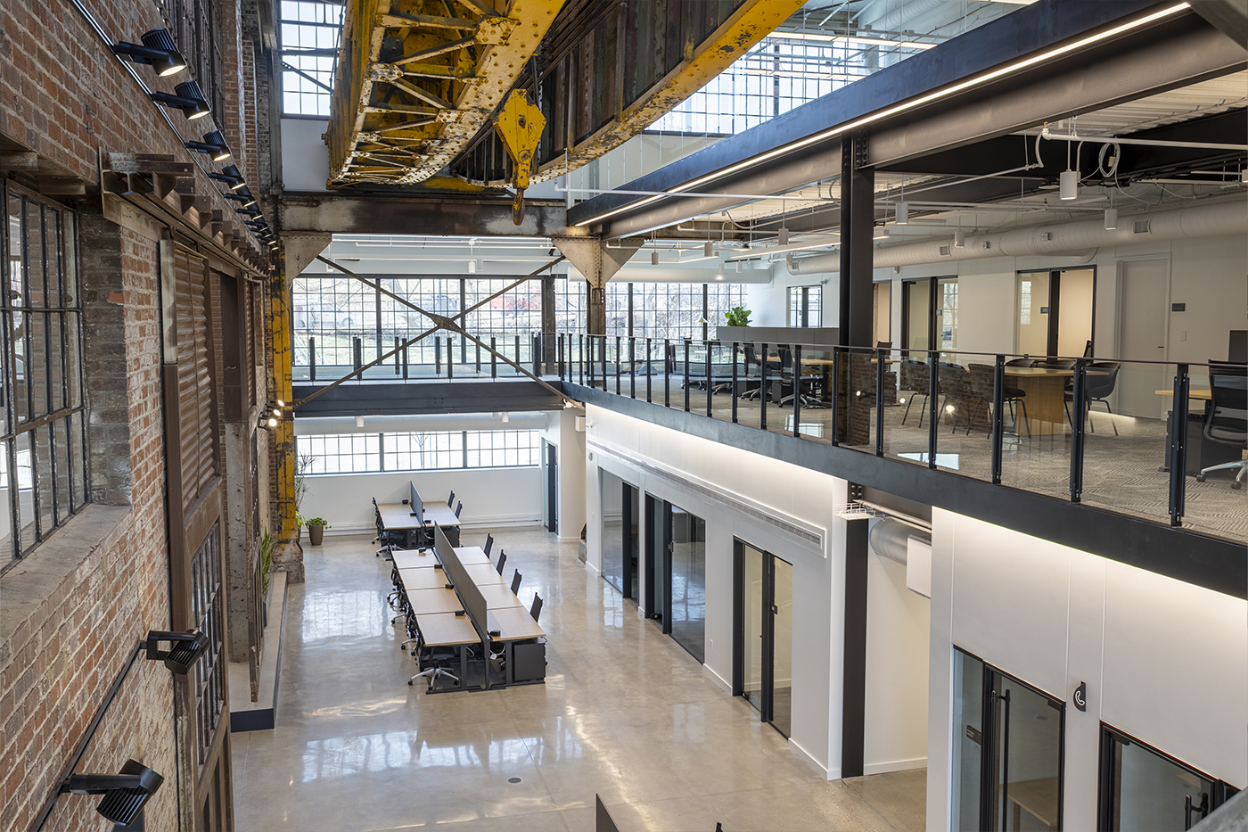 Modern industrial office space with exposed brick walls, large windows, open workstations, and a mezzanine level with glass railing.