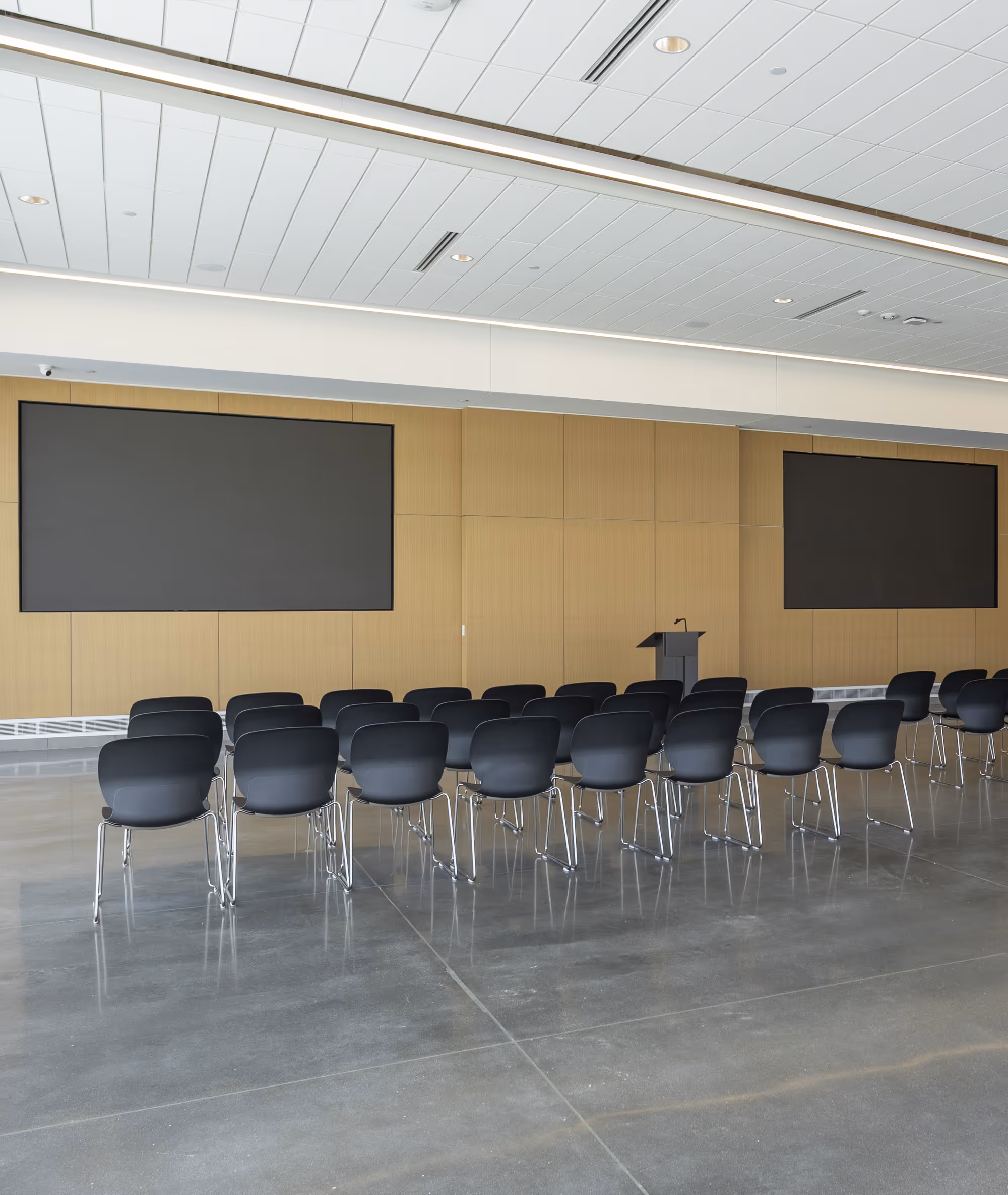 Modern conference room with rows of black chairs facing two large blank screens and a podium against a wood-paneled wall.