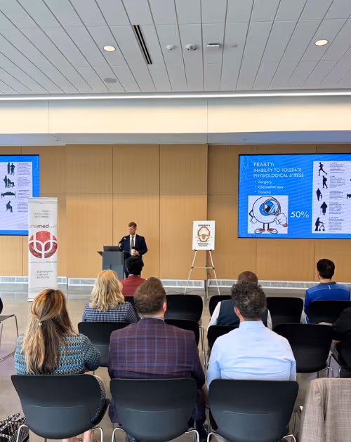 A man in a suit giving a lecture in a conference room with audience members seated and a large screen showing a presentation on physiological stress and frailty.
