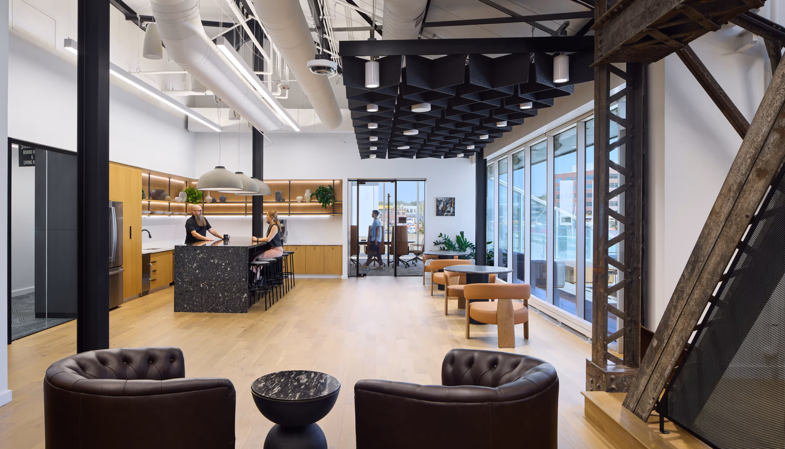 Modern office lounge with brown leather chairs, a kitchen island with people sitting, wooden floors, and large windows overlooking a city.