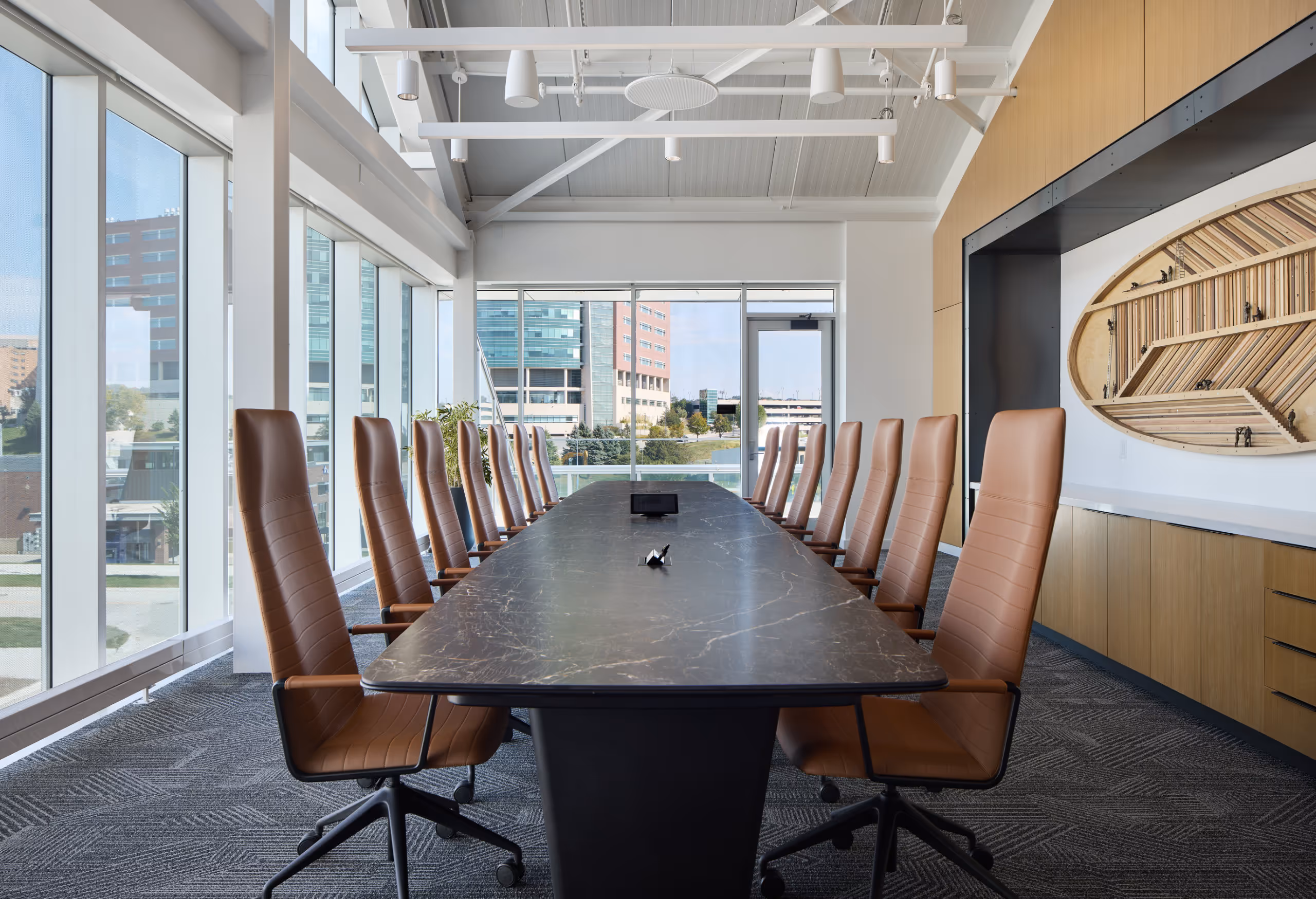 Modern boardroom with a long black table, fourteen brown leather chairs, large windows showing city buildings, and a wooden art installation on the wall.