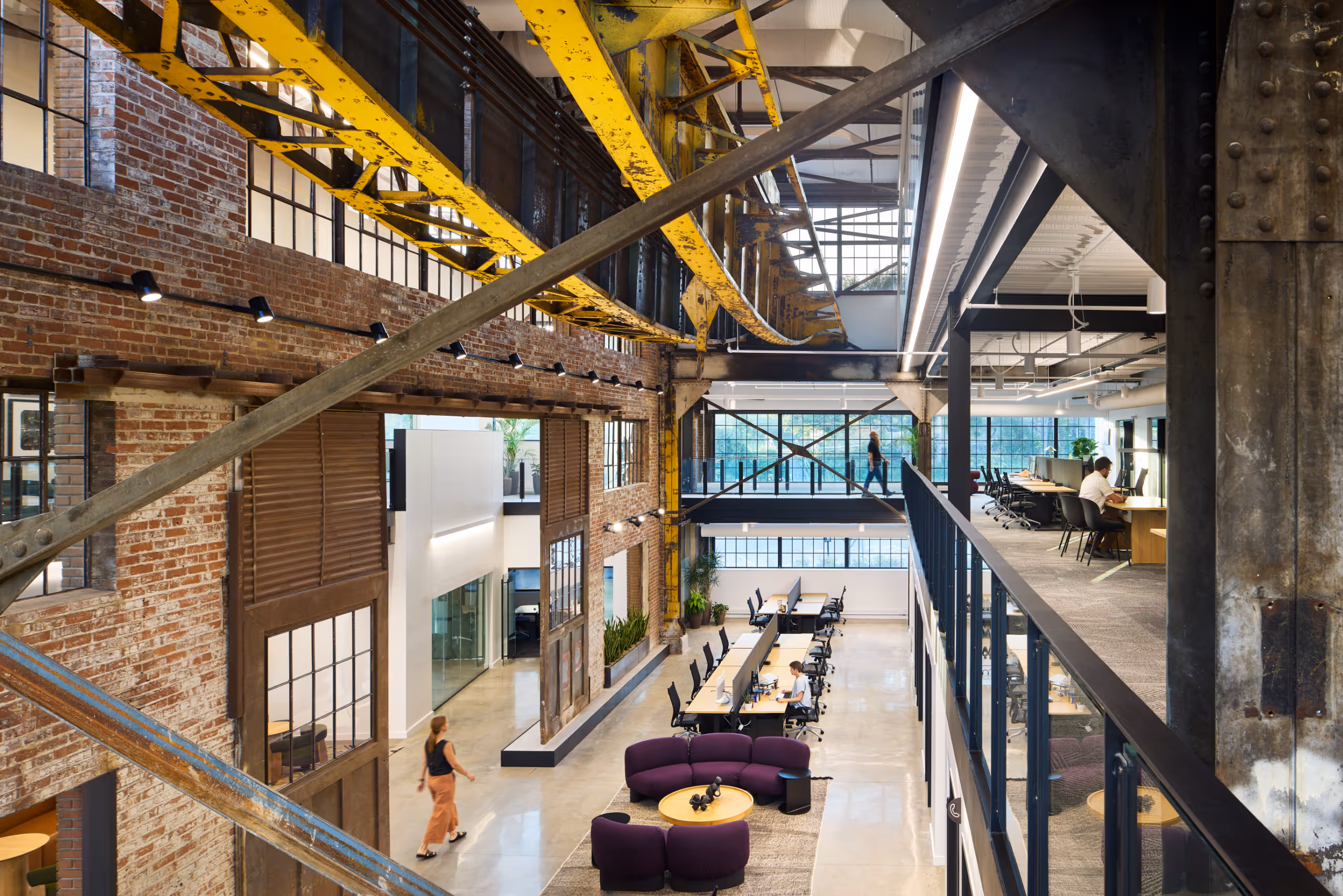 Modern industrial coworking space with exposed brick walls, yellow overhead crane beams, open desks, purple lounge seating, and people working and walking.
