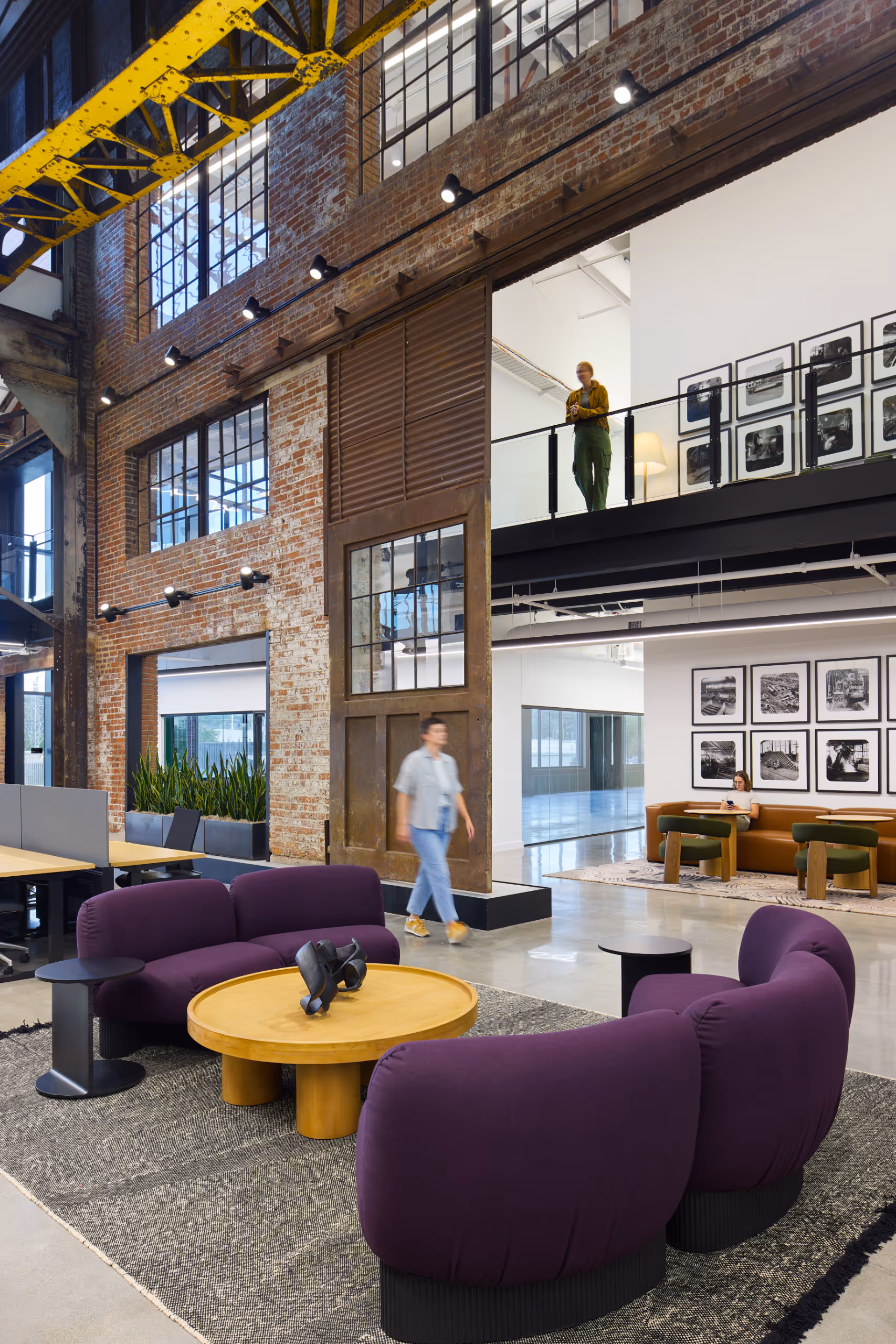 Modern coworking lounge with purple curved sofas around a wooden coffee table under industrial yellow girders and large windows.