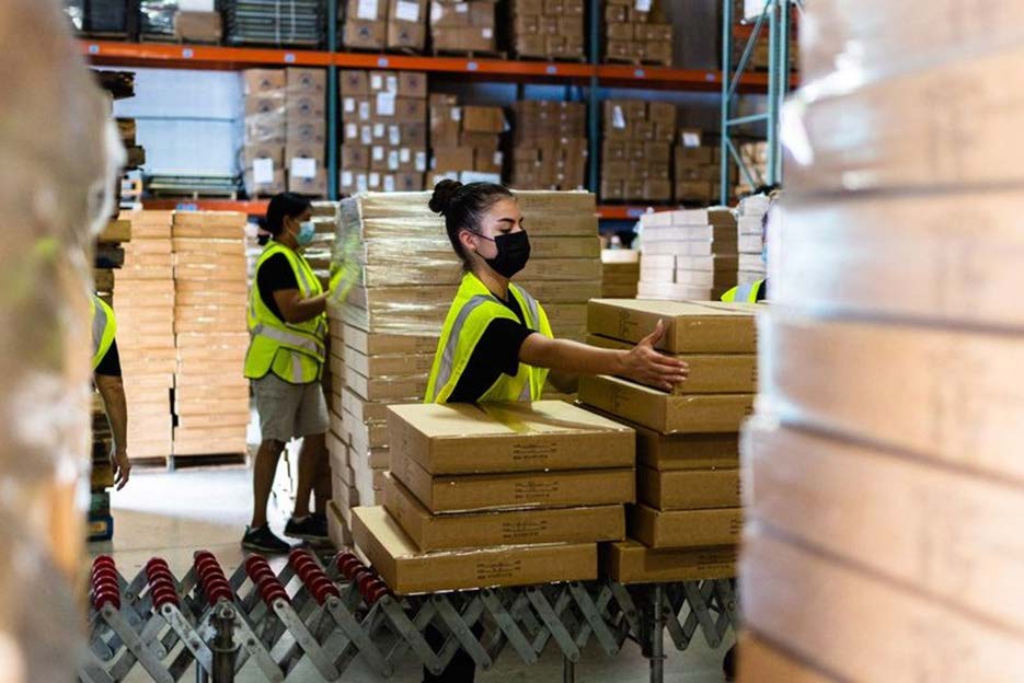 A woman wearing a mask and a yellow vest is working in a warehouse.