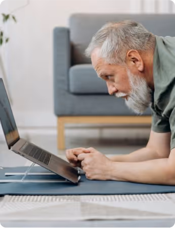 Older man with gray beard working on laptop while lying on floor mat