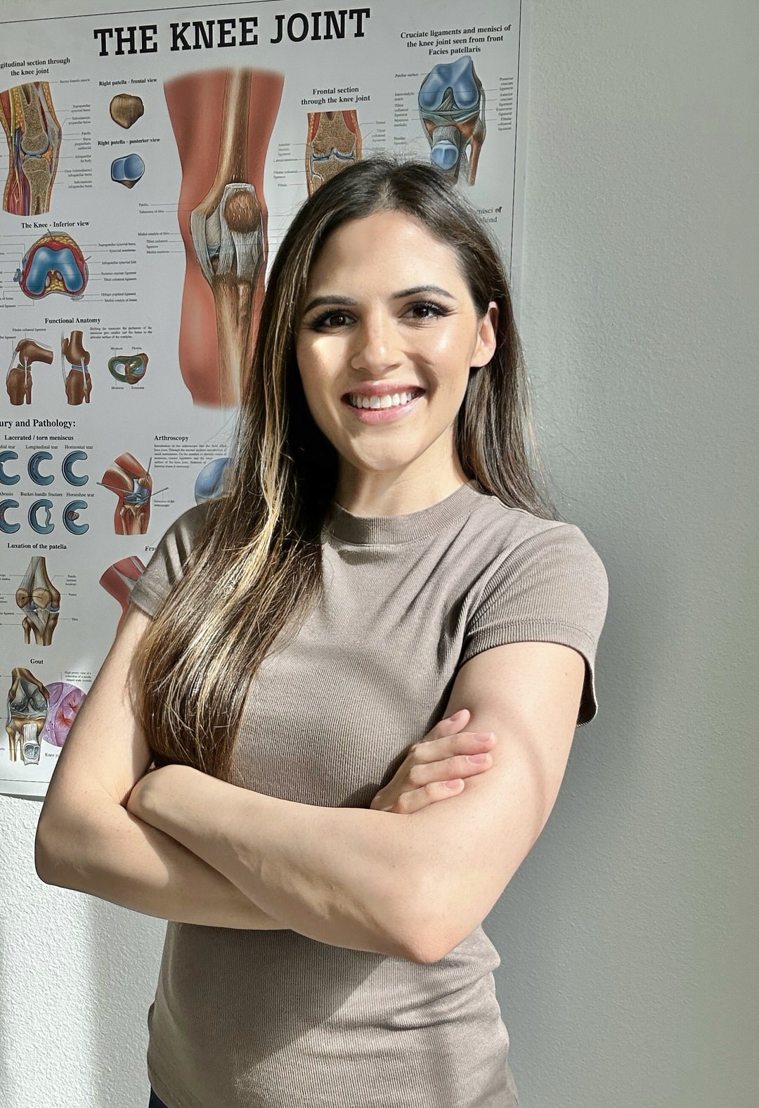 Smiling medical professional standing in front of detailed knee joint anatomy poster