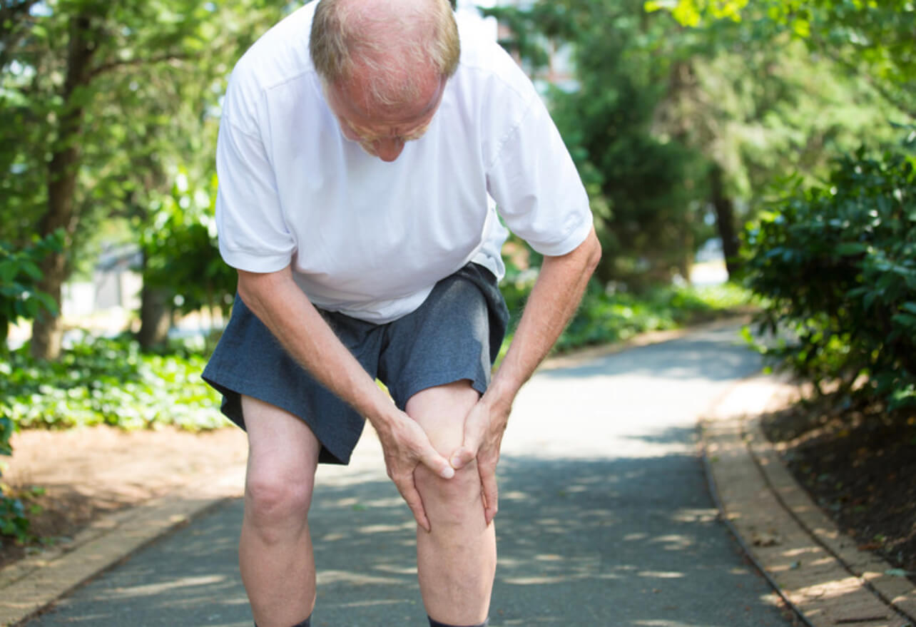 Older person holding knee in pain during outdoor walk on tree-lined path