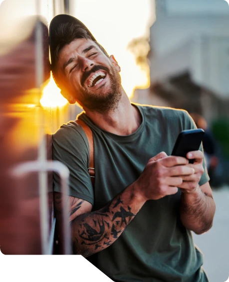 Foto de um rapaz de boné e roupa casual, rindo muito e feliz ao olhar seus resultados na tela de seu celular.