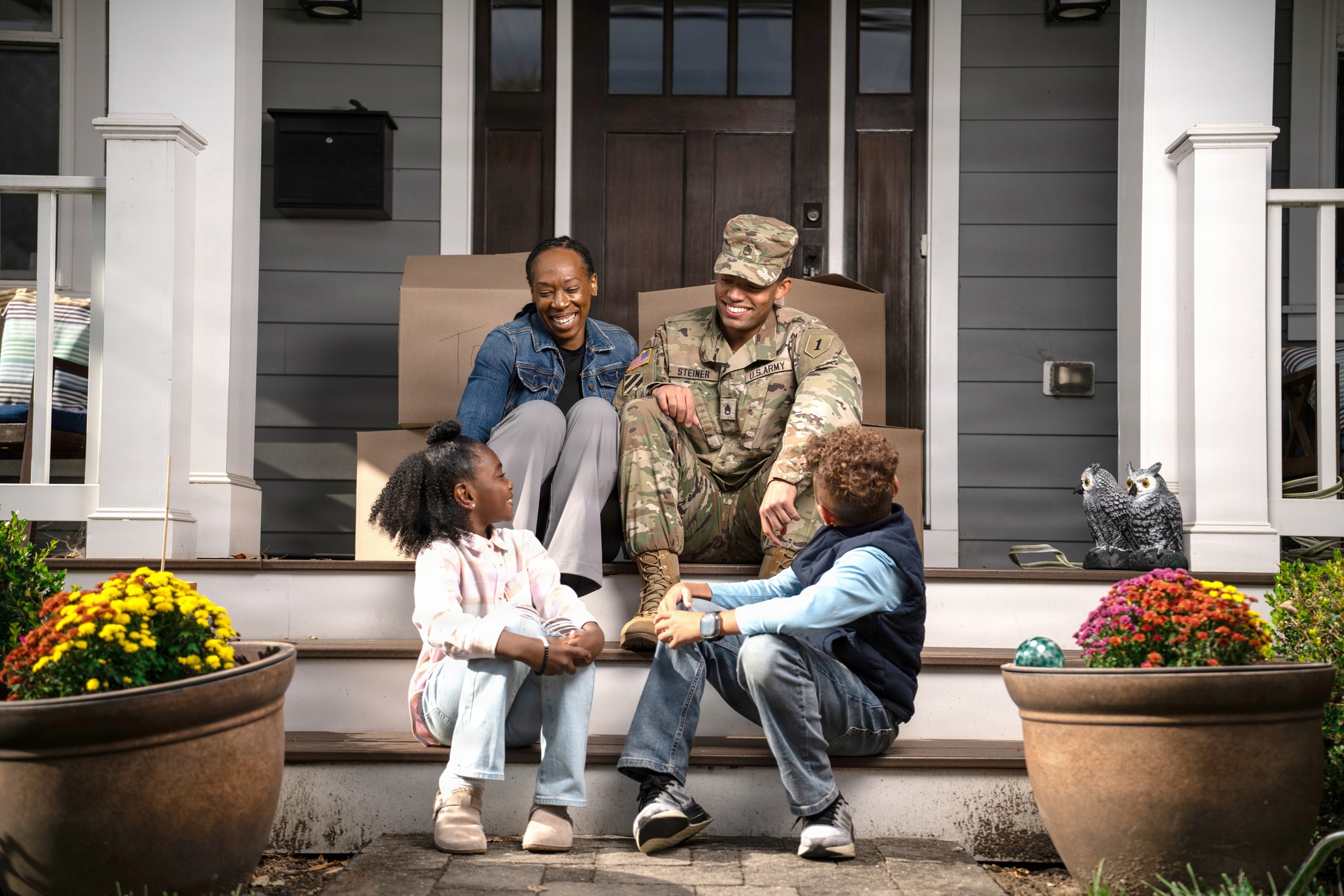 A Soldier and his family sit on the front-porch steps of their new home with moving boxes behind them.