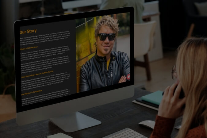 Person with blonde hair and glasses sitting at a desk, looking at a computer screen displaying a webpage titled 'Our Story' with a photo of a man wearing sunglasses and a leather jacket.