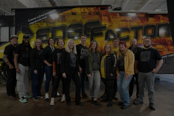 A group of 13 people standing indoors in front of a large vehicle branded with 'EDGE FACTOR'.