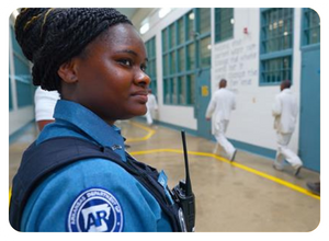 Female correctional officer in uniform standing inside a prison corridor with two inmates walking in the background.