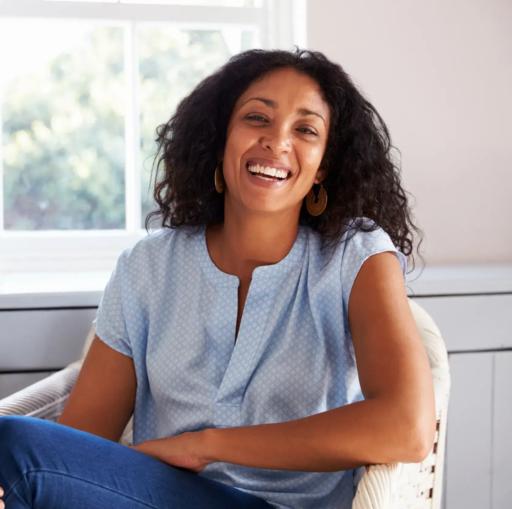A woman sits smiling in a chair near a window, wearing a blue blouse.