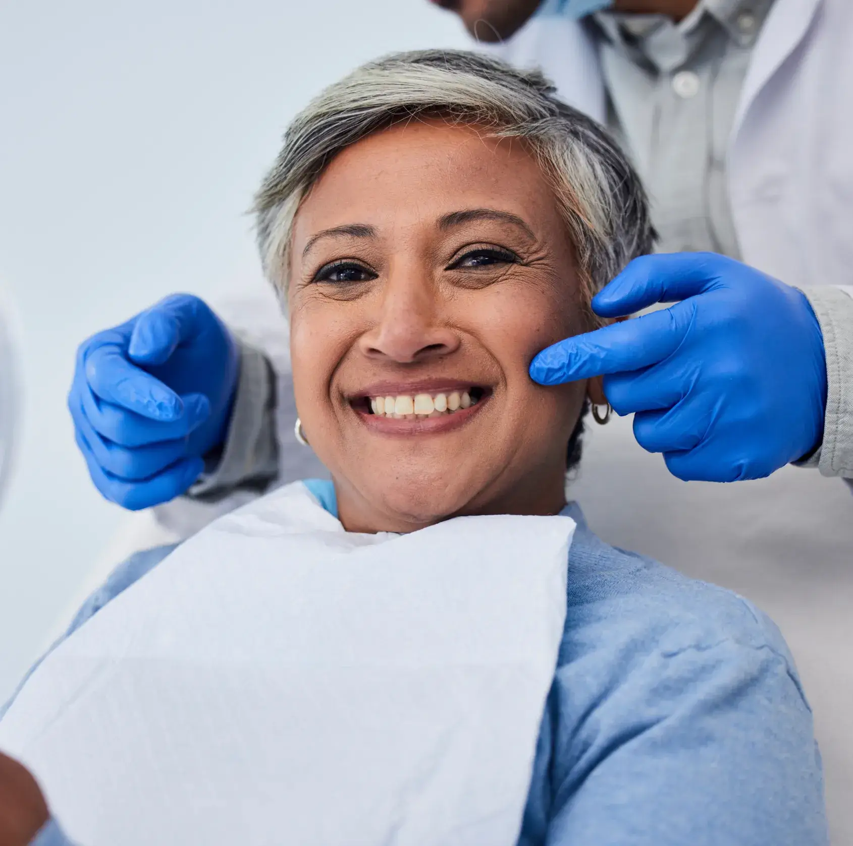 Smiling woman sitting in a dental chair while a dentist adjusts her mouth with gloved hands.