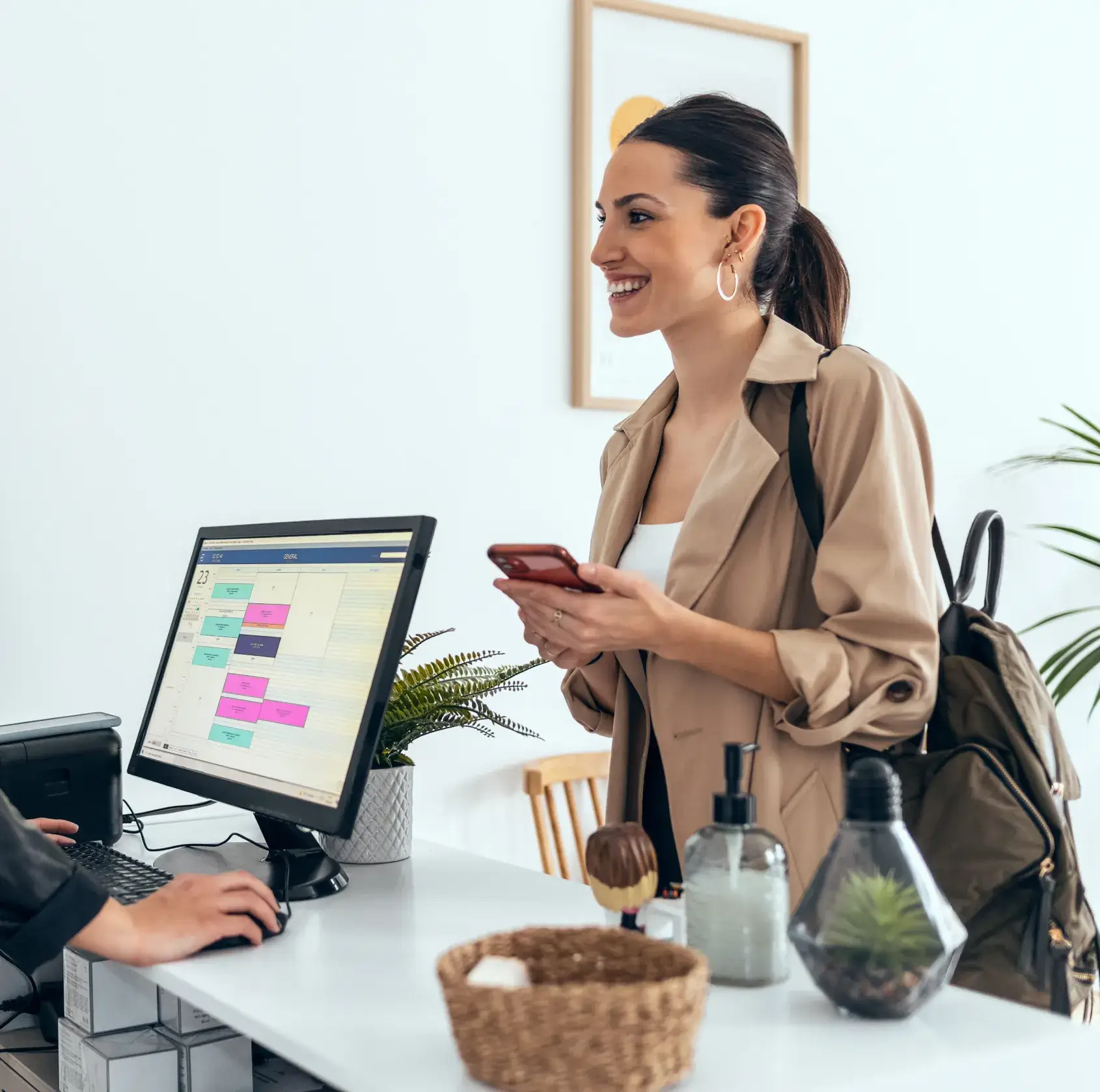 Woman standing at a desk smiling, holding her phone, while another person uses a computer.