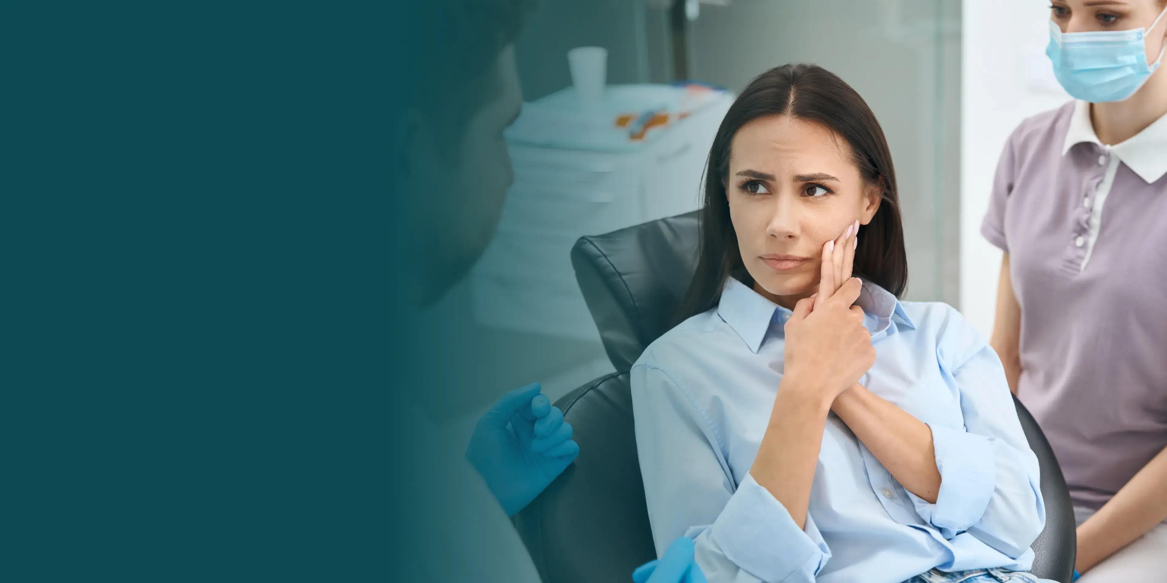 A woman in a dentist chair touches her cheek, looking concerned while listening to a dentist.