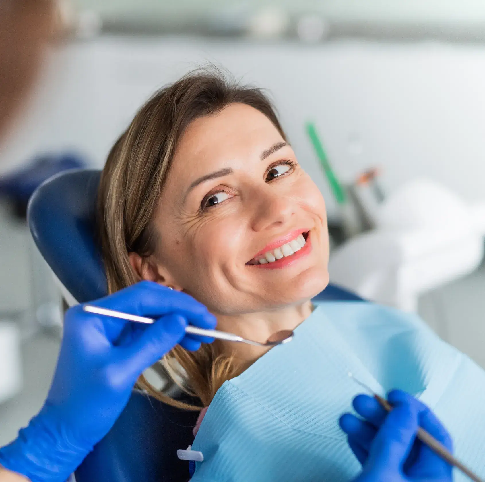A woman smiles while sitting in a dental chair as a dentist holds instruments nearby.