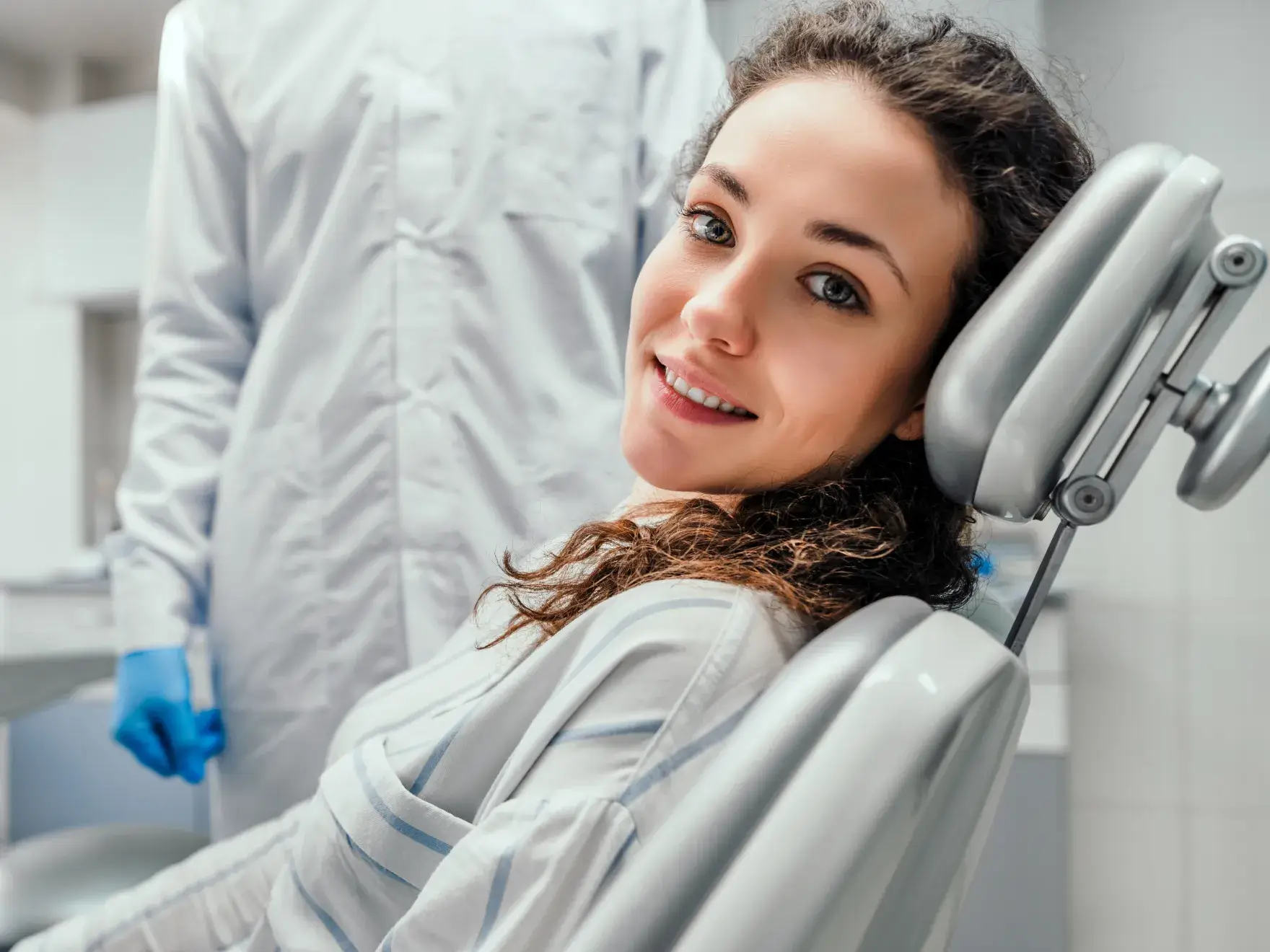 A woman sitting in a dental chair smiles while a dentist wearing a white coat stands nearby.