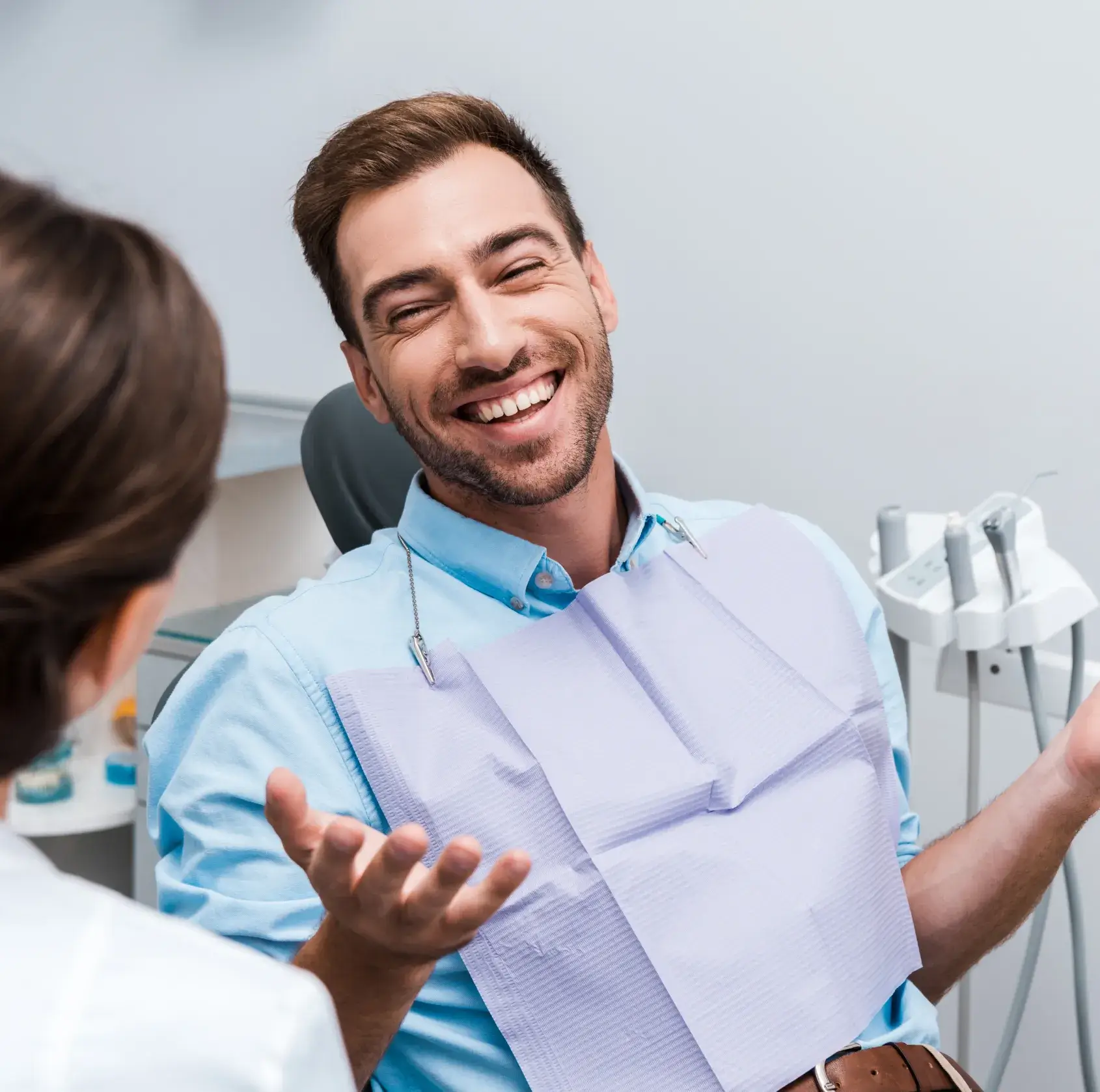 A man with a bib smiles and gestures while sitting in a dental chair, facing a dentist.