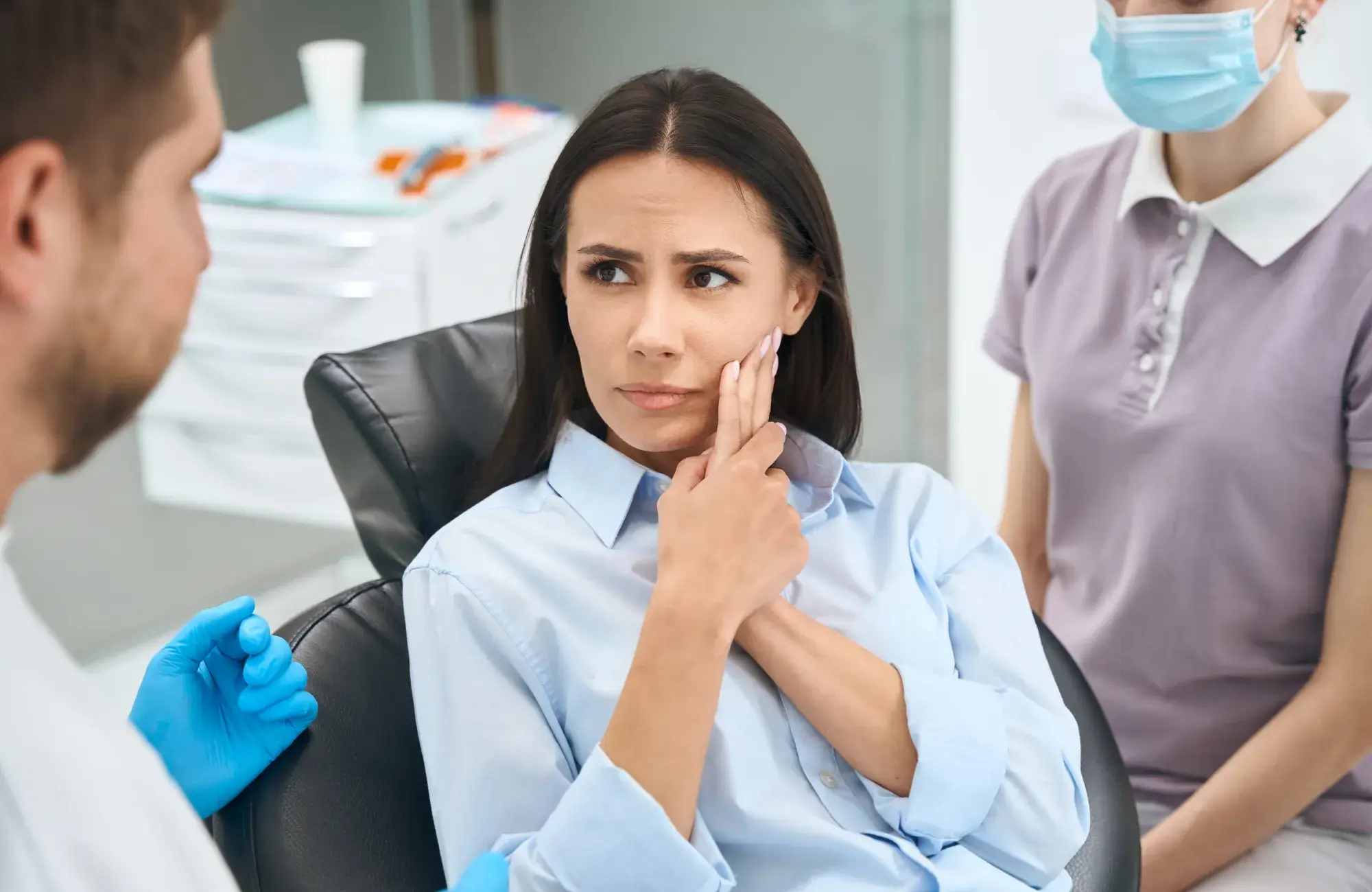 Woman in a dentist's chair touching her cheek, looking concerned. Dentist and assistant nearby.
