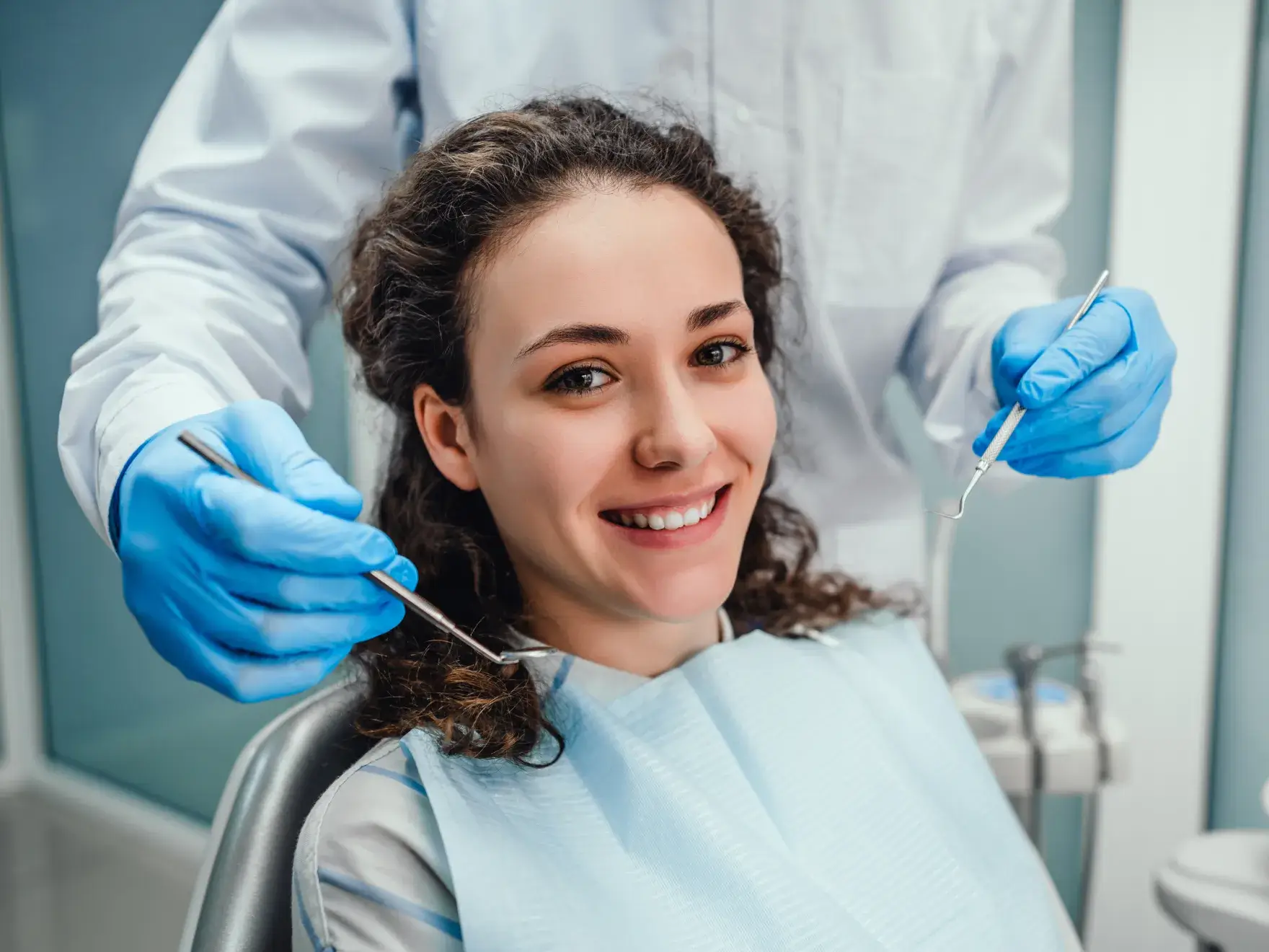 Woman sitting in a dental chair, smiling, while a dentist holds dental tools near her mouth.