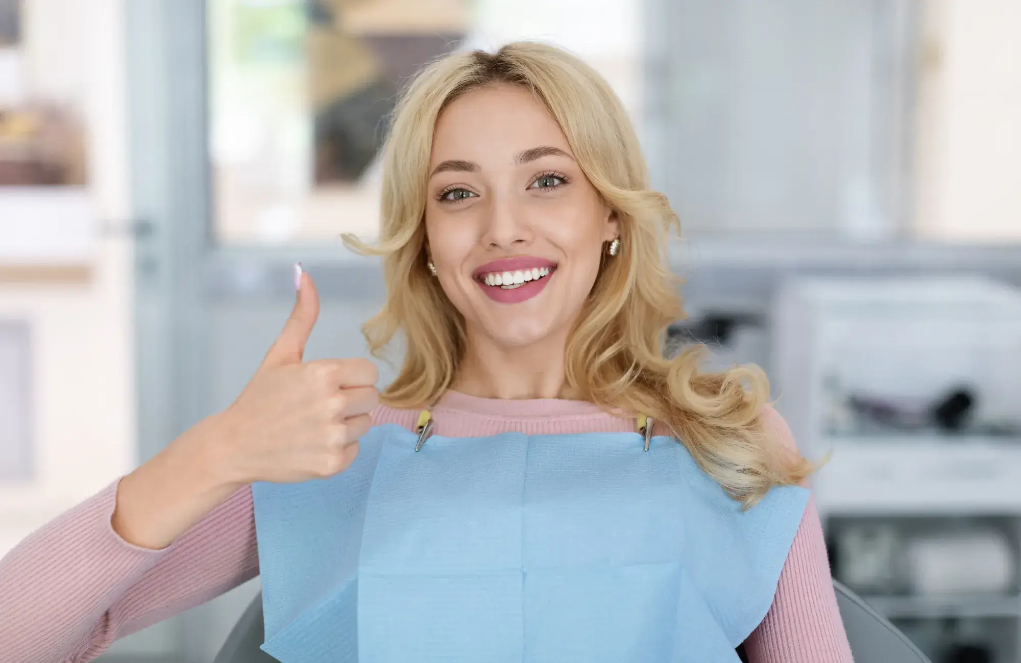 A woman sitting in a dental chair gives a thumbs-up while smiling.