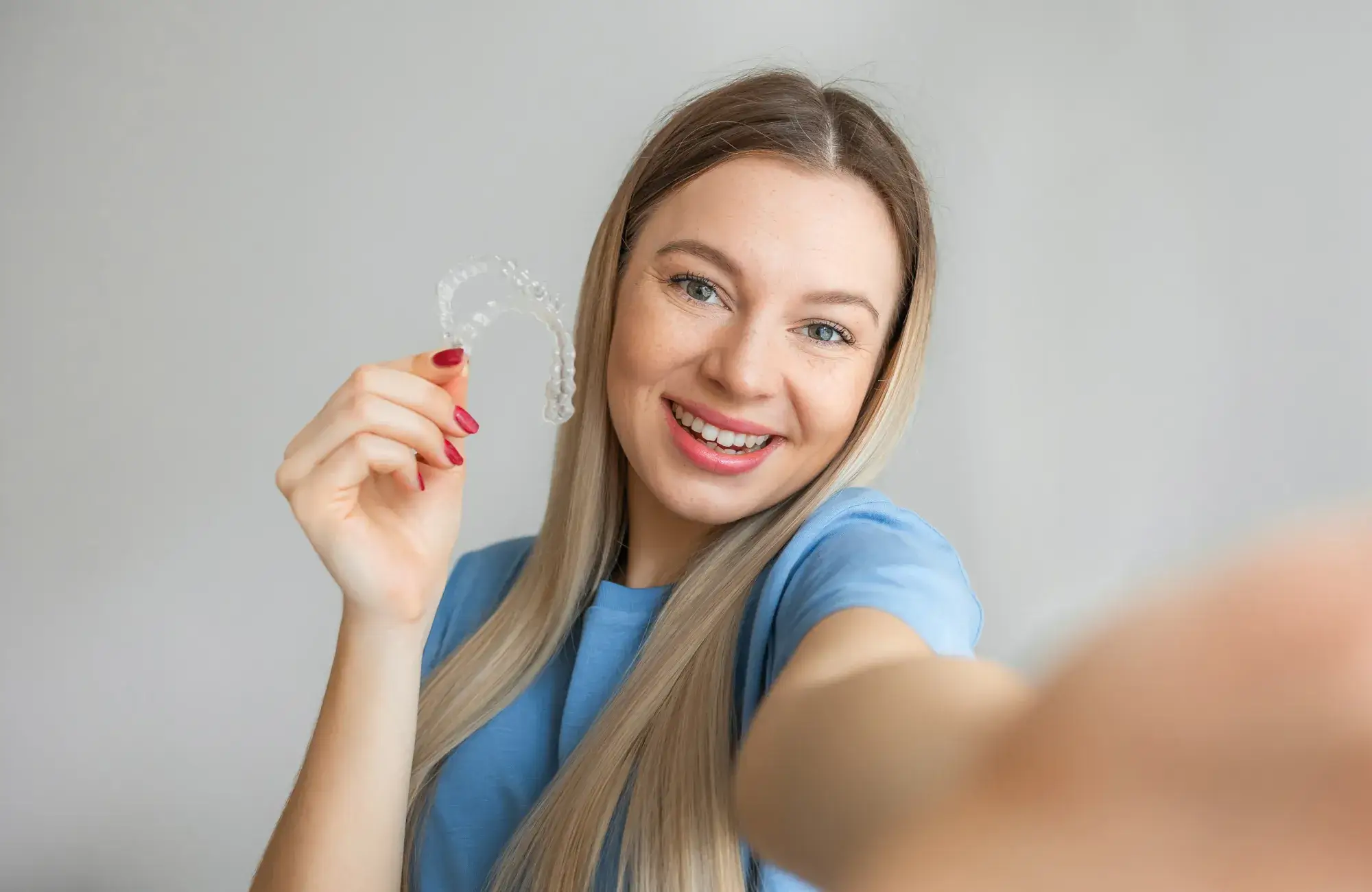 A person smiling and holding a clear dental aligner in their hand.