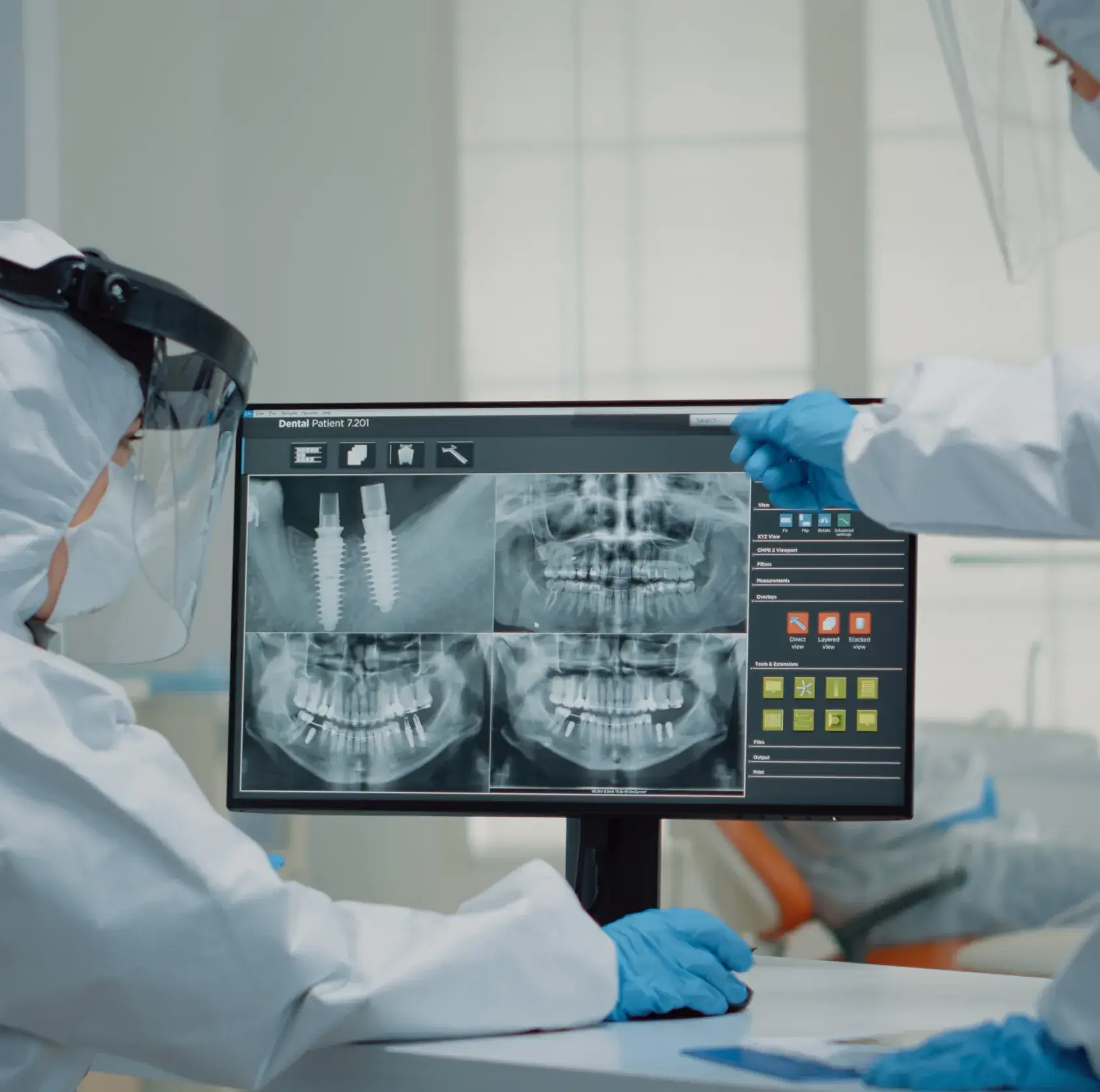 Two dentists in protective gear examine dental X-rays on a computer monitor in an office.