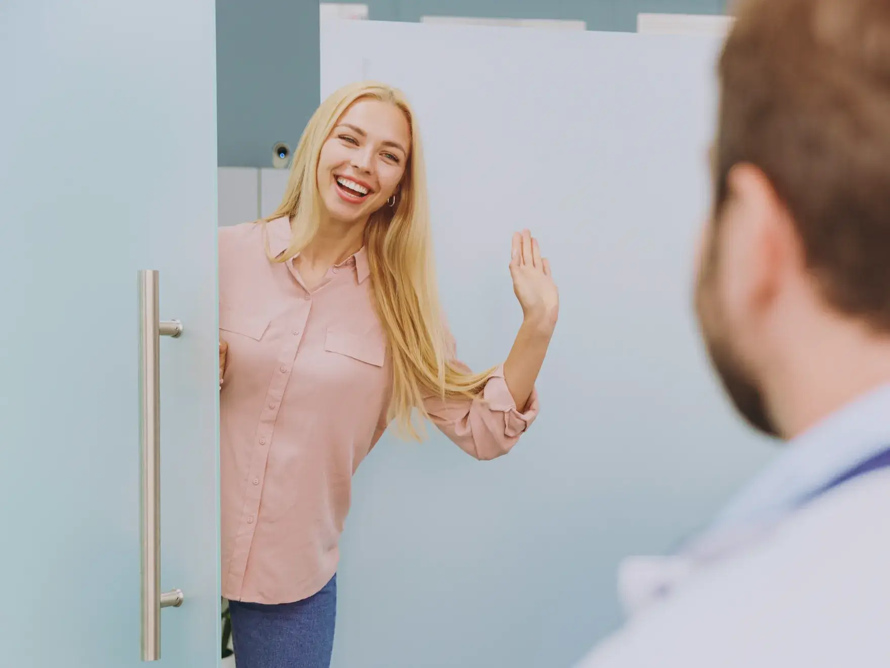 A woman with long blonde hair waves and smiles at a person in front of her.