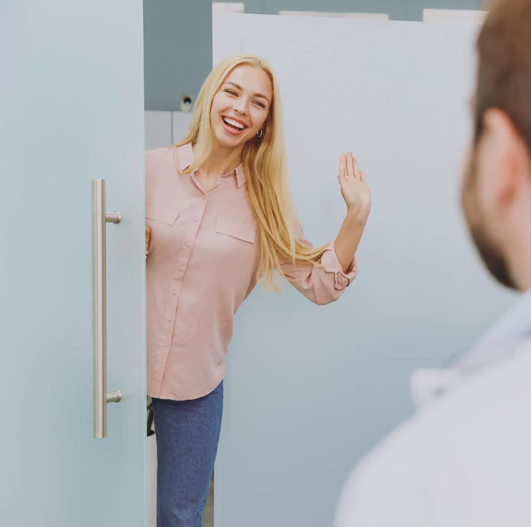 Woman in a pink shirt waves and smiles while entering a room through an open door.
