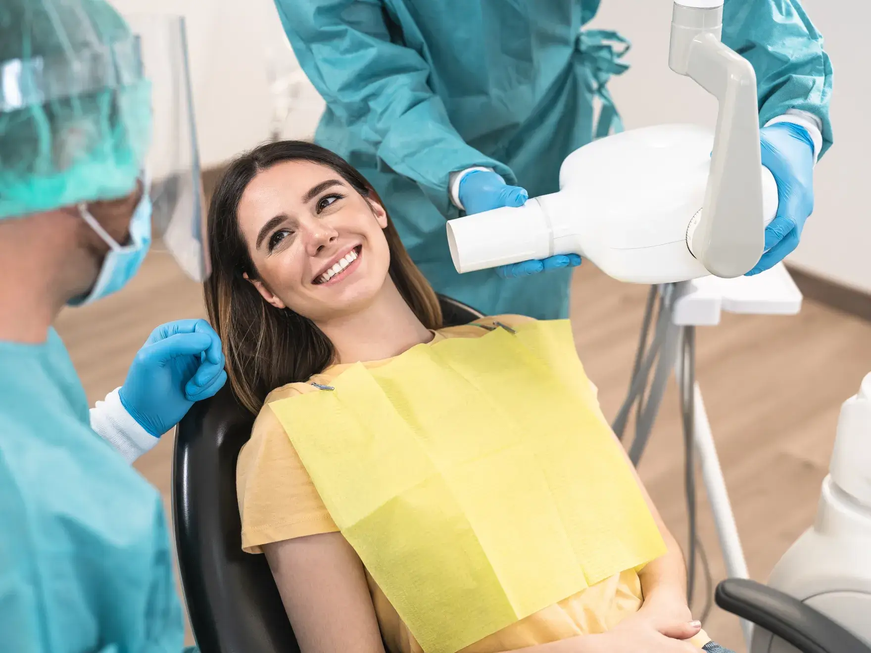 A woman smiles while sitting in a dental chair, surrounded by dentists preparing equipment.