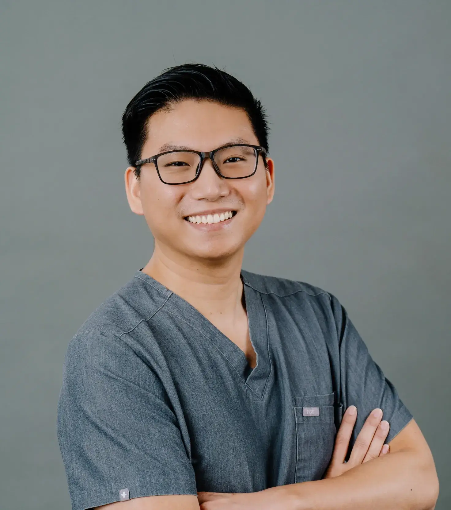 Smiling person in blue scrubs with arms crossed against a plain background.