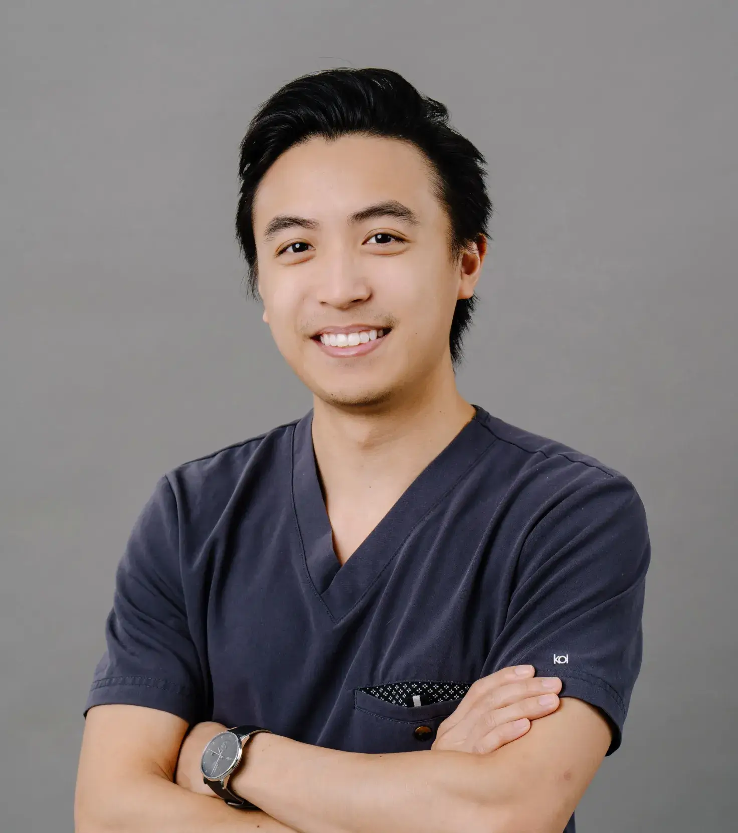 A person in a dark scrubs top smiles with crossed arms against a plain background.