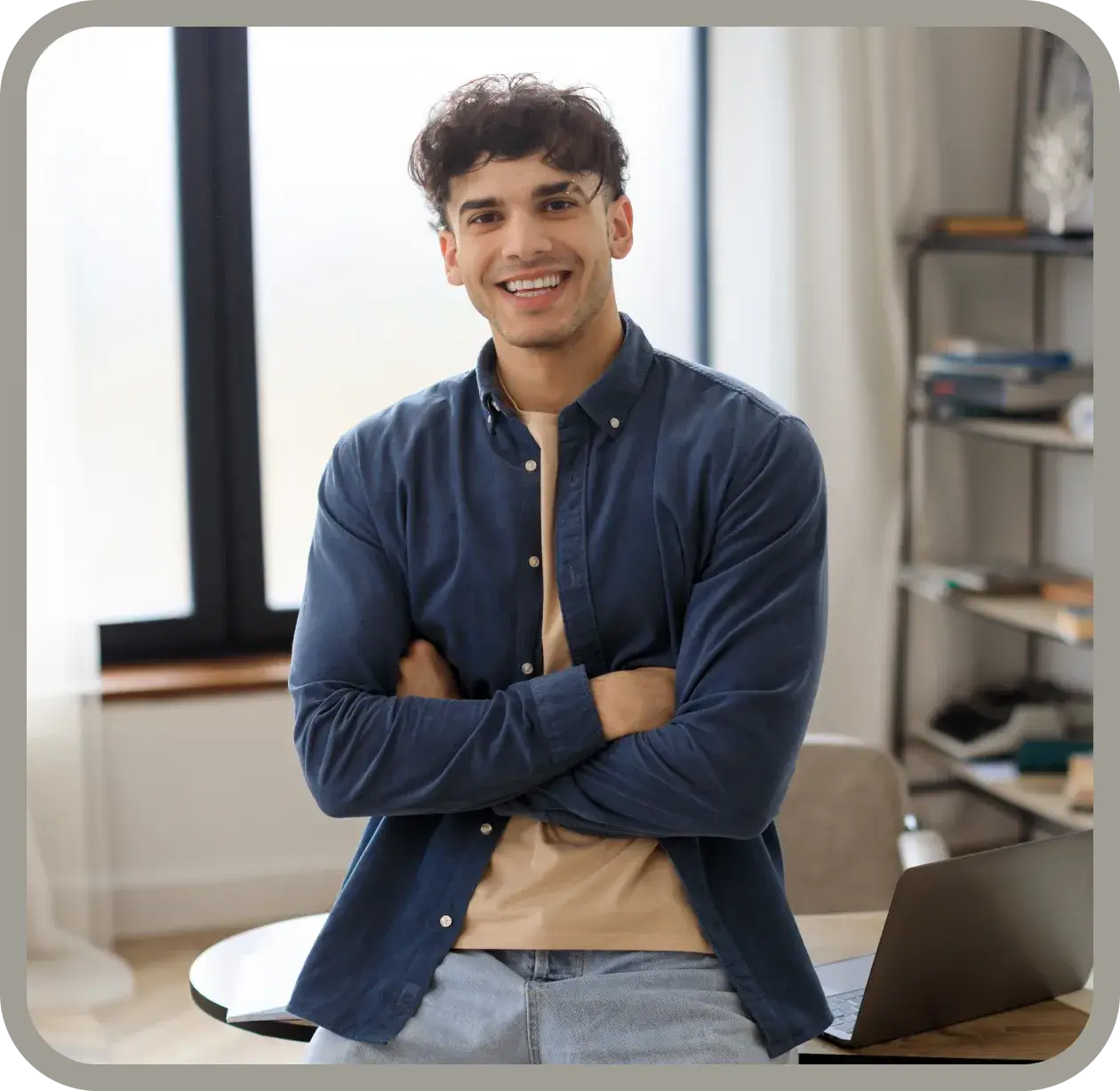 A man stands indoors, smiling with arms crossed, next to a table with a laptop.