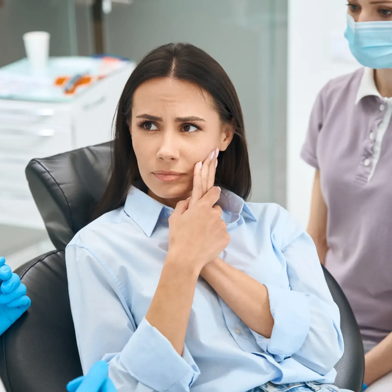 A woman sitting in a dental chair touches her cheek, looking concerned. Two masked dental professionals nearby.