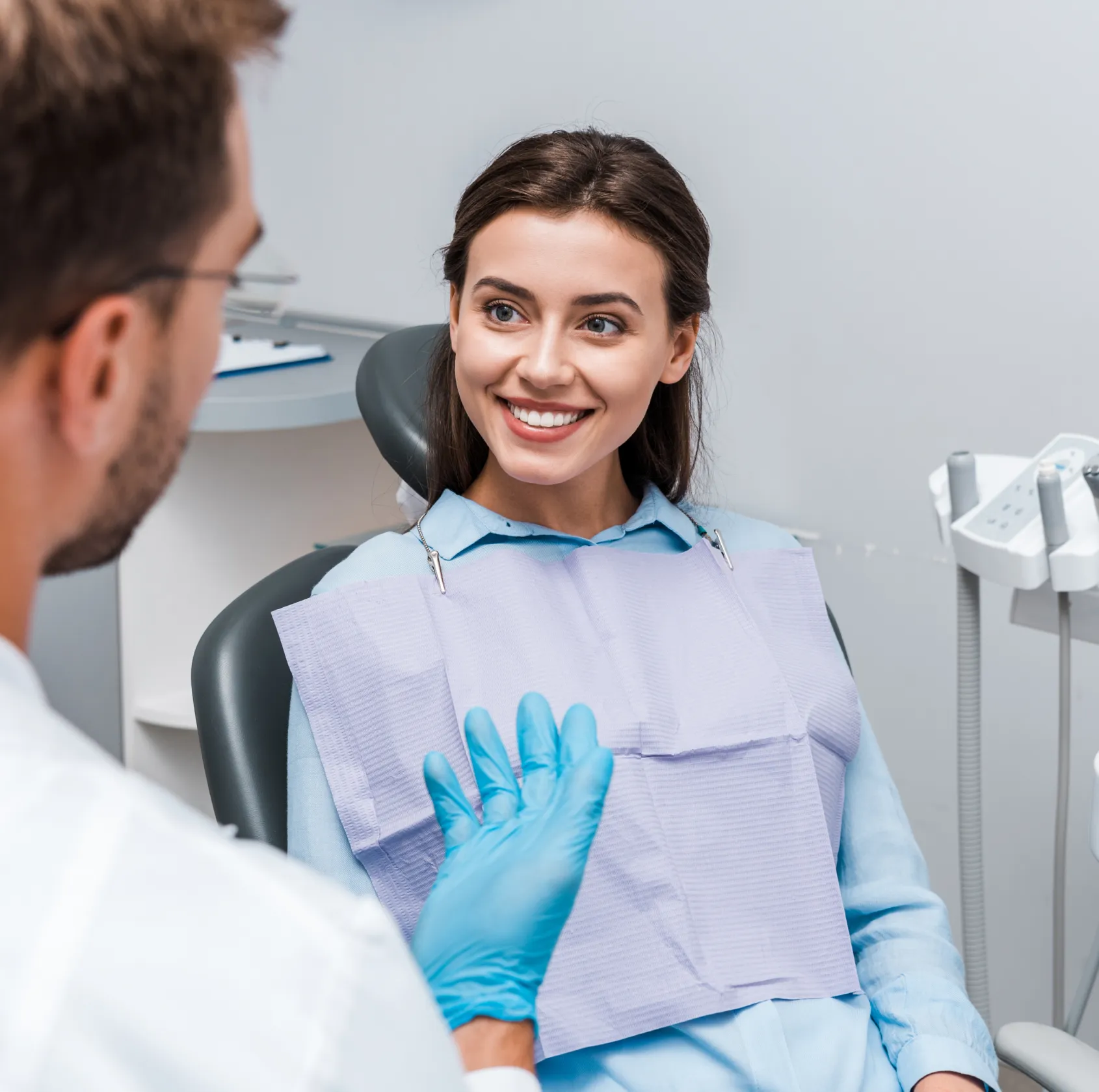 Dentist in gloves talks to a patient sitting in a dental chair, smiling.