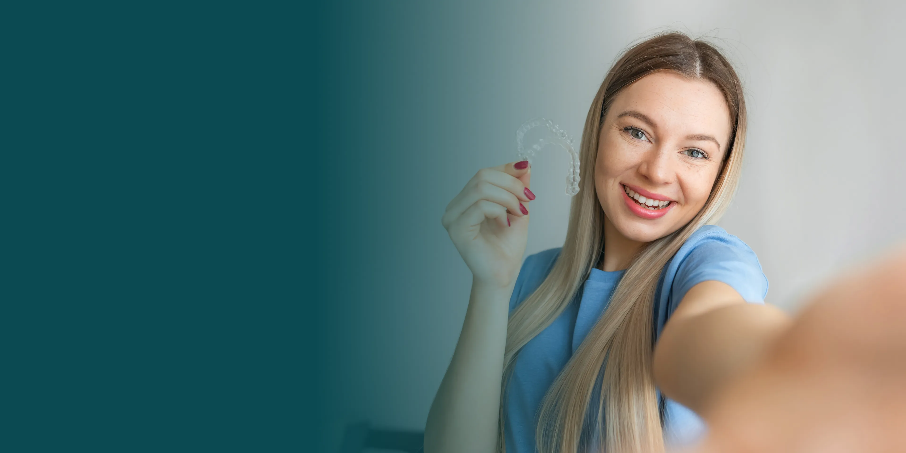 Woman smiling and holding a dental retainer while taking a selfie.