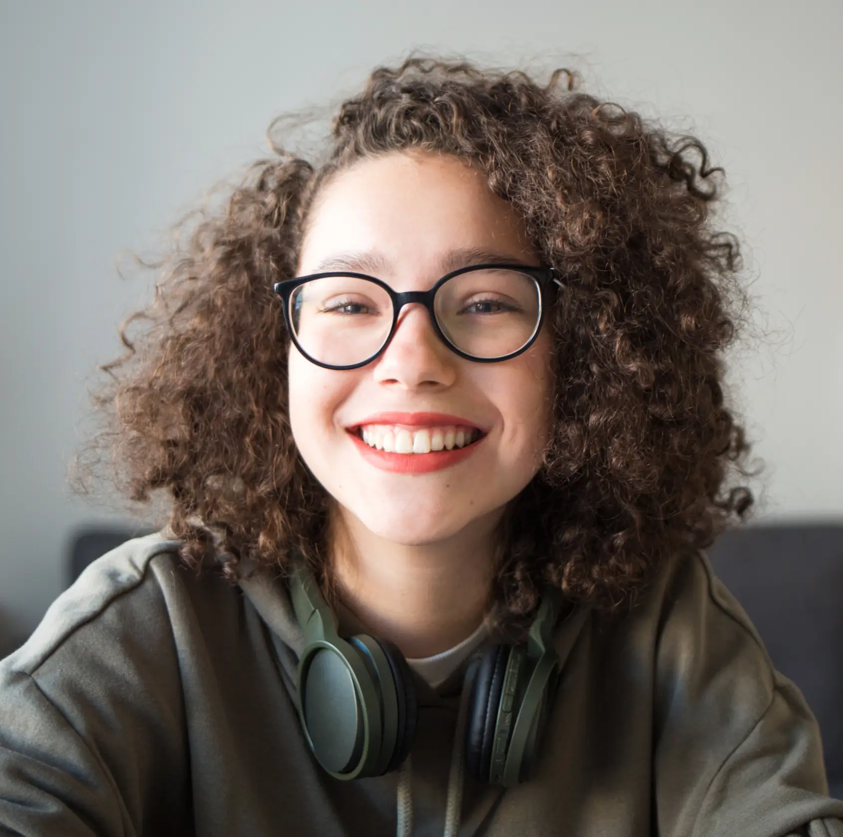 Young person with curly hair and glasses smiling, wearing headphones around their neck.