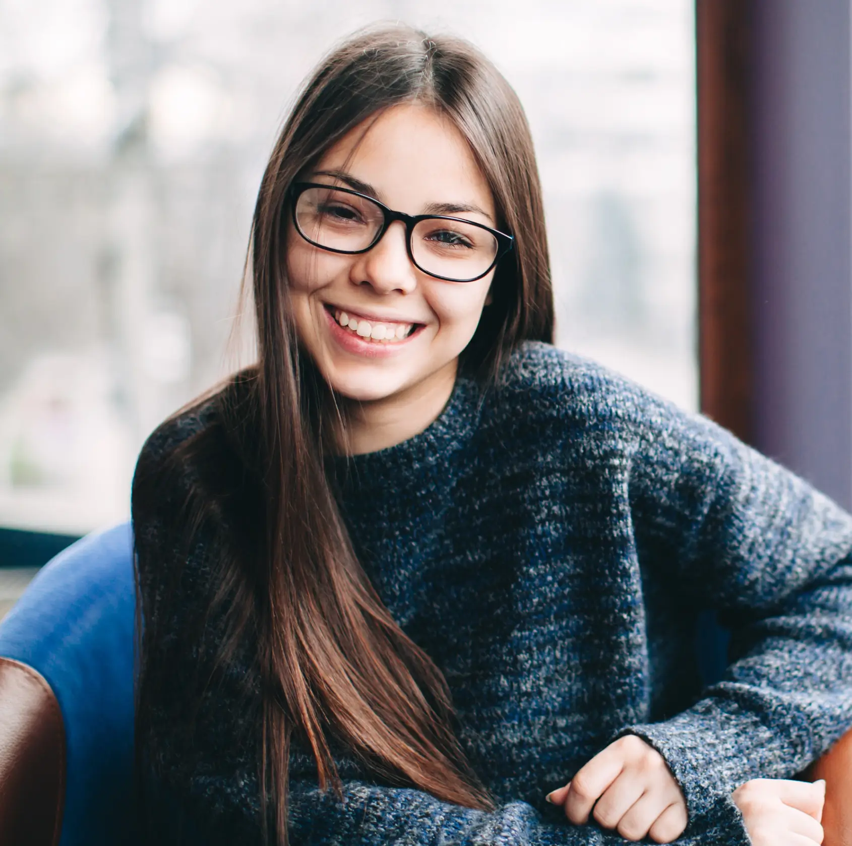 A young woman with long hair and glasses smiles while sitting indoors.