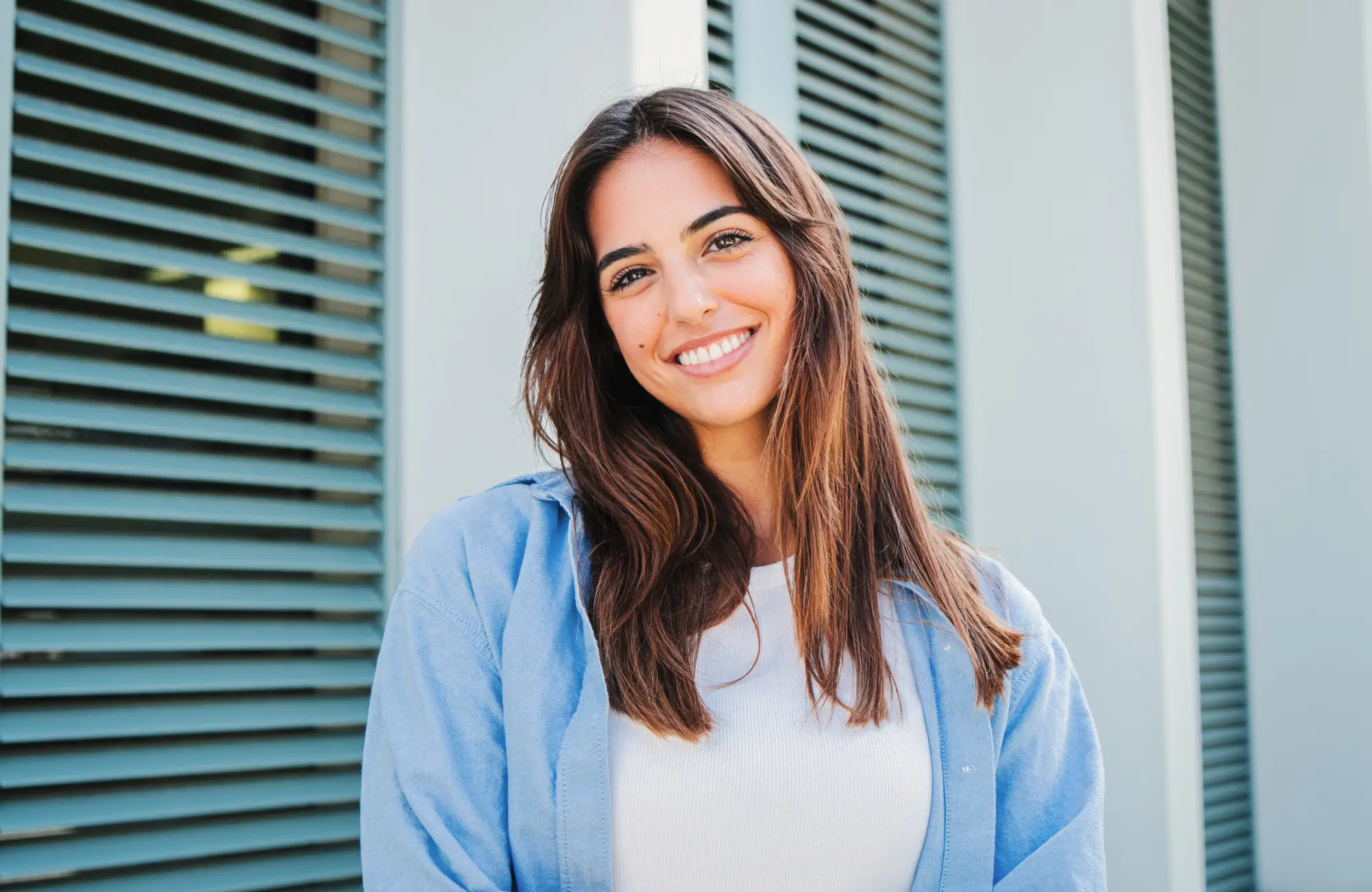 A woman with long brown hair smiles warmly, standing in front of blue shutters.