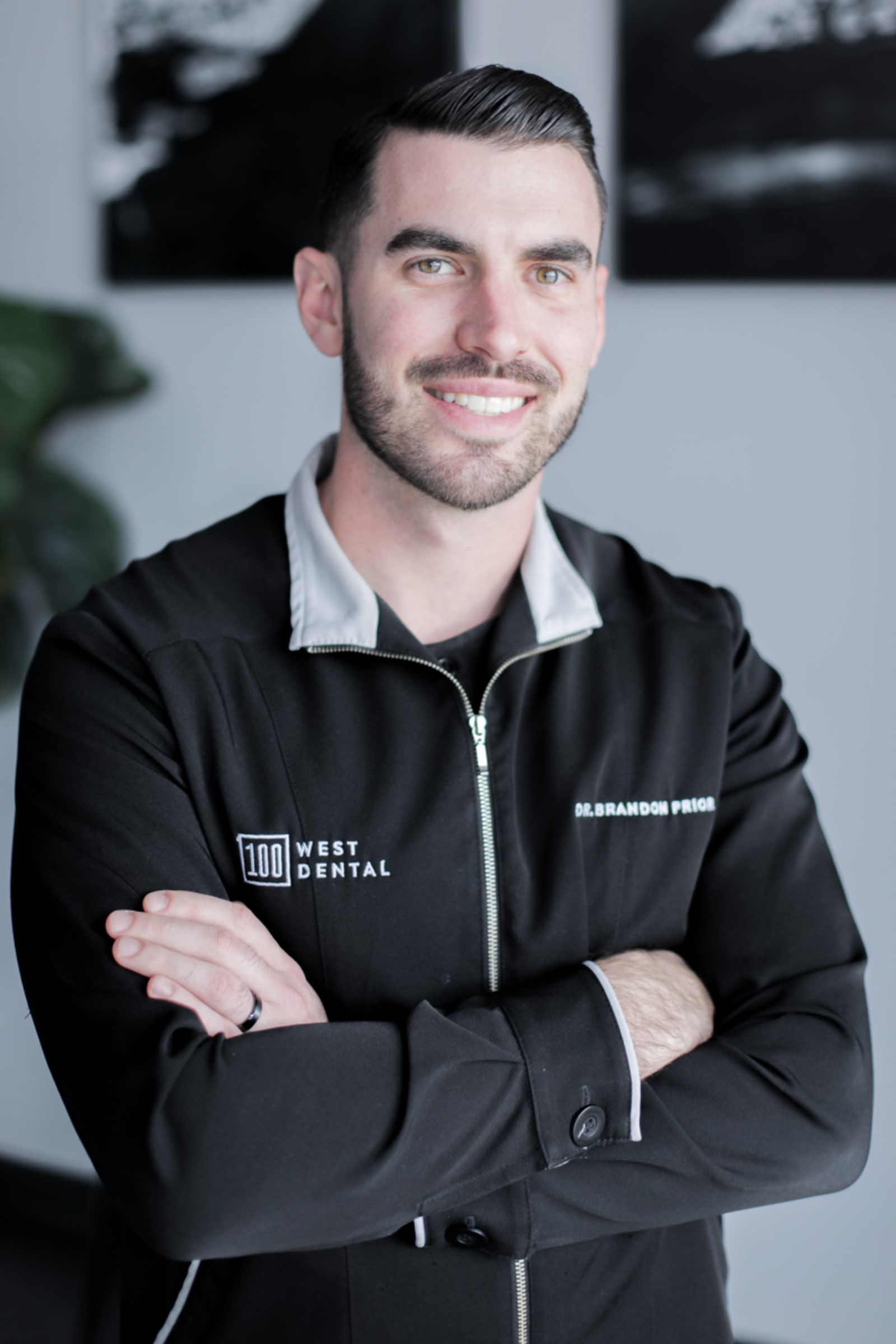 Professional headshot of a male dentist in black 100 West Dental branded jacket smiling at camera.