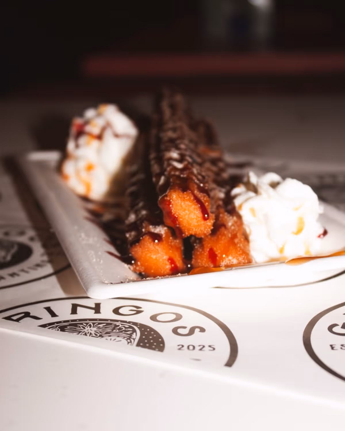 Three churros drizzled with chocolate sauce on a white rectangular plate, served with dollops of whipped cream on the sides.