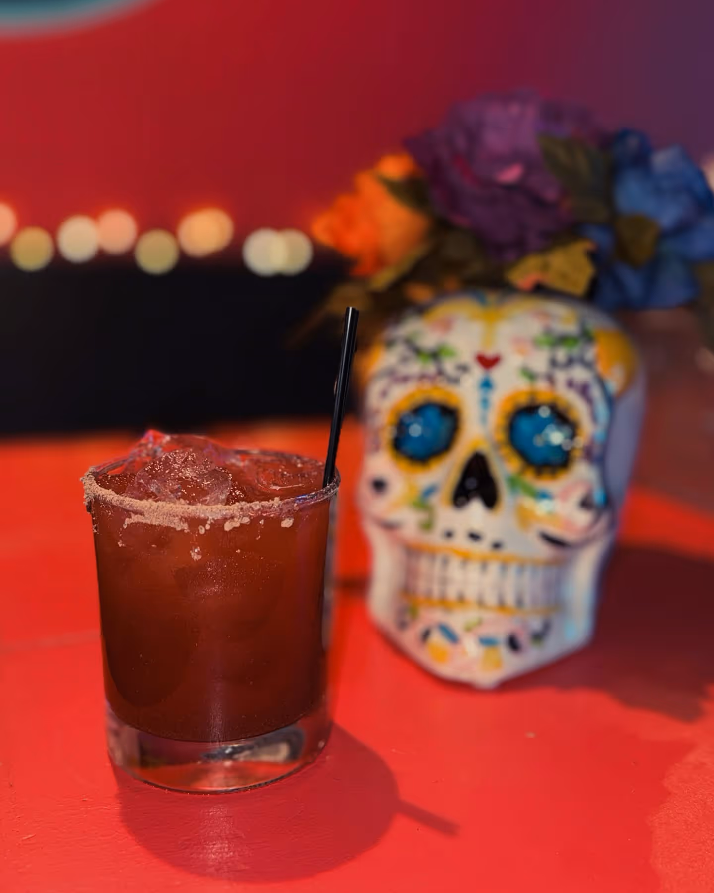 Glass of red cocktail with salted rim and ice on a red surface, with a colorful decorative skull with flowers blurred in the background.