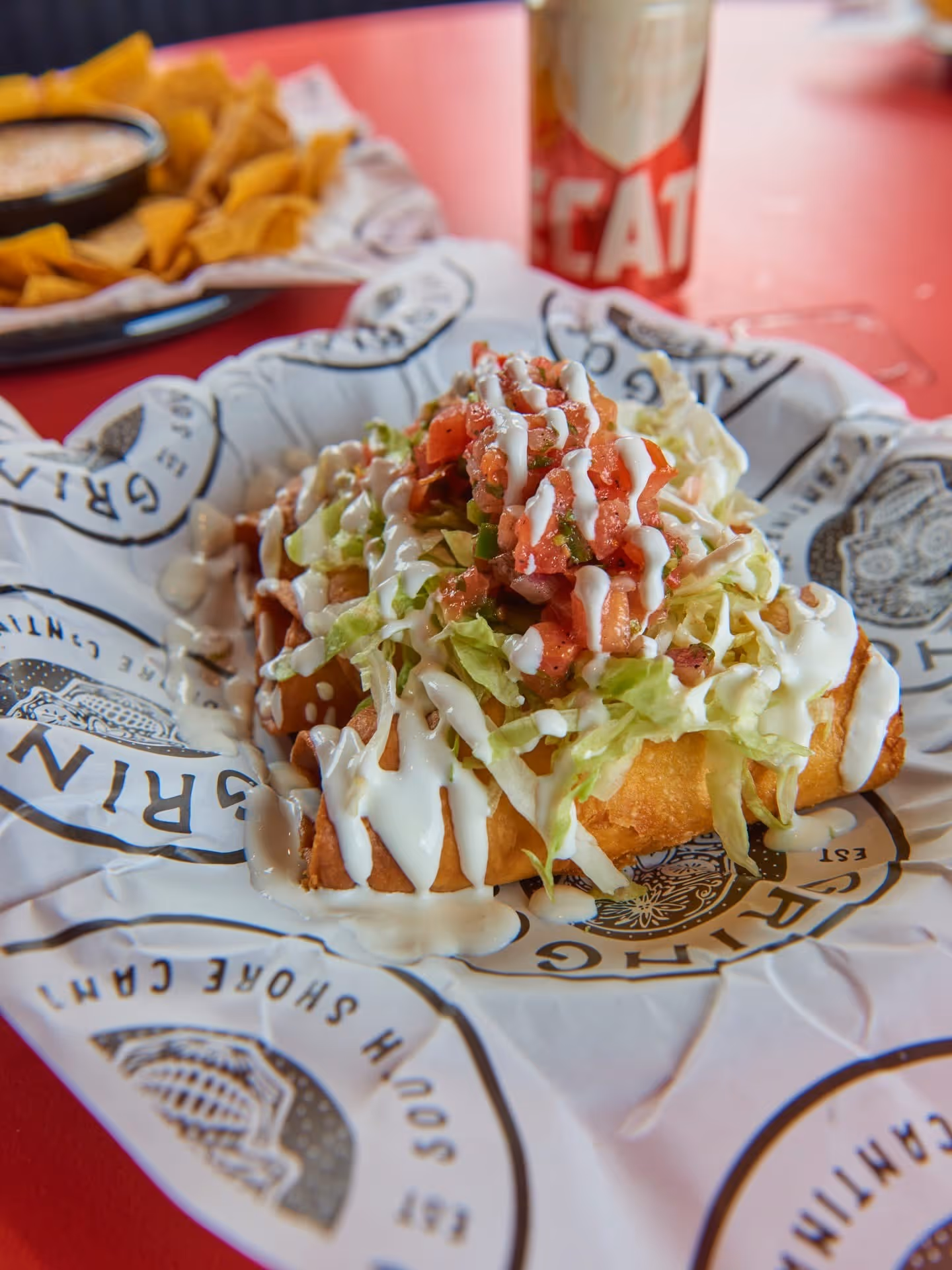 Fried taco topped with shredded lettuce, diced tomatoes, and drizzled with white sauce on branded paper.